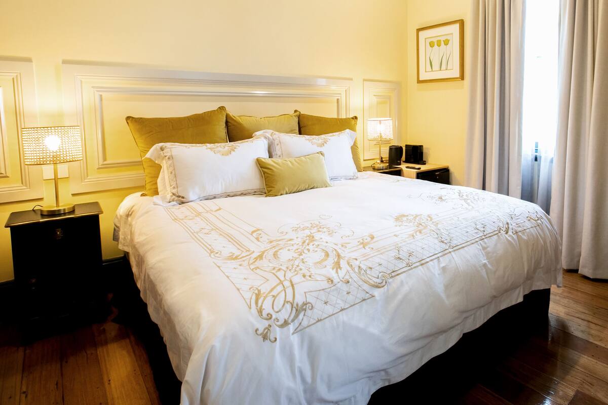 A neatly made bed with a white bedspread and gold embroidery, flanked by two nightstands with lamps, in a room with wooden flooring and a large window with curtains.