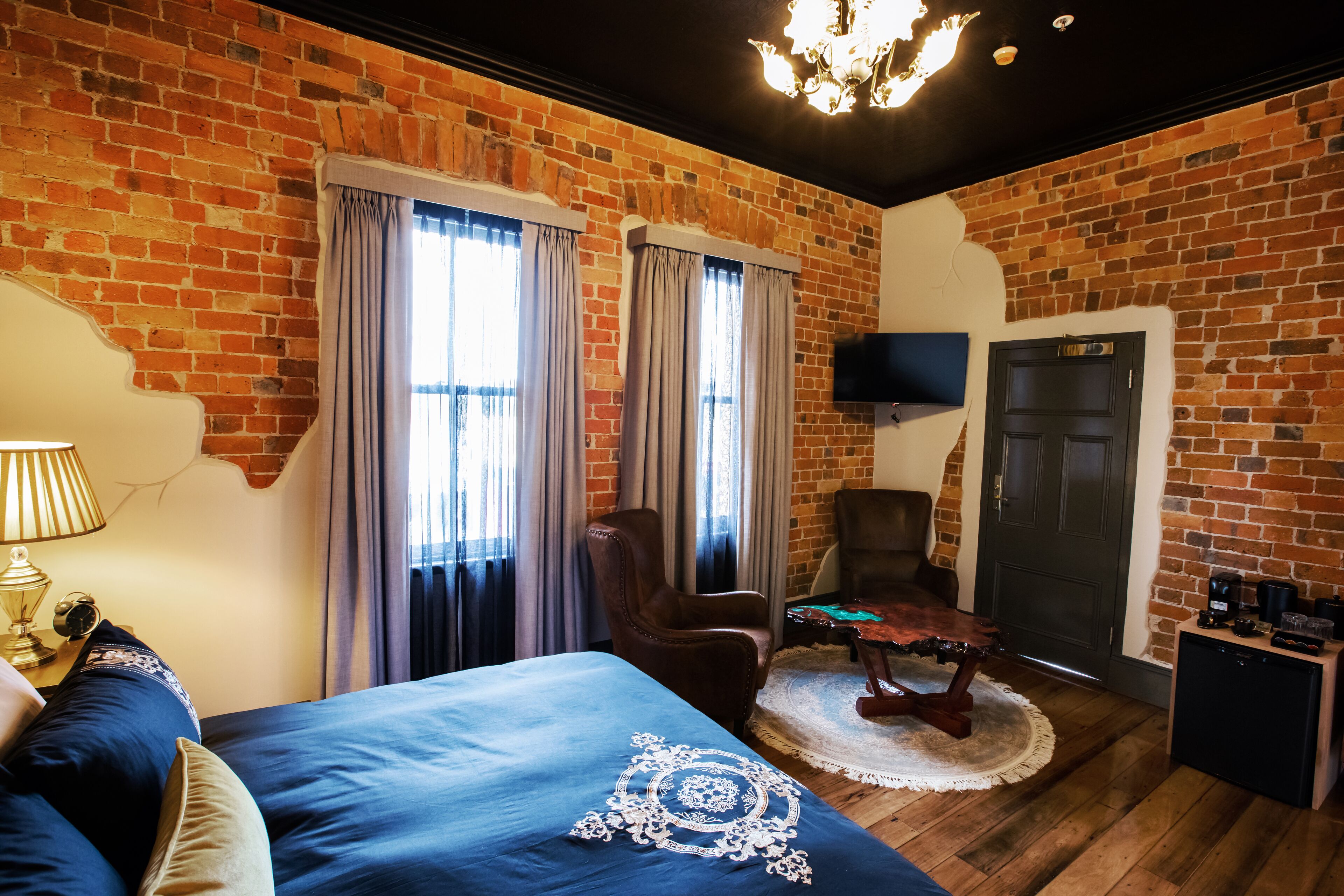 Ein Zimmer mit Backsteinwand, einem Bett mit blauer Bettdecke, einem braunen Ledersessel, einem Holztisch und einem an der Wand montierten Fernseher.