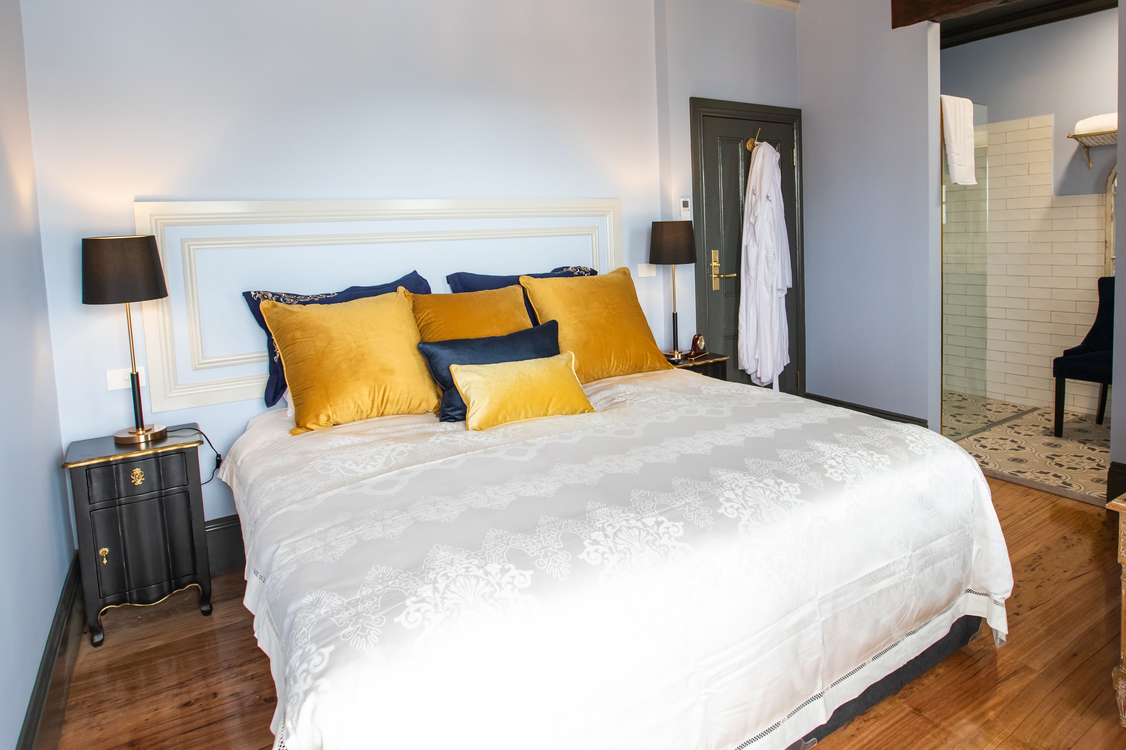 A neatly made bed with a white and gold bedspread, two nightstands with lamps, and a door with a towel hanging on it.