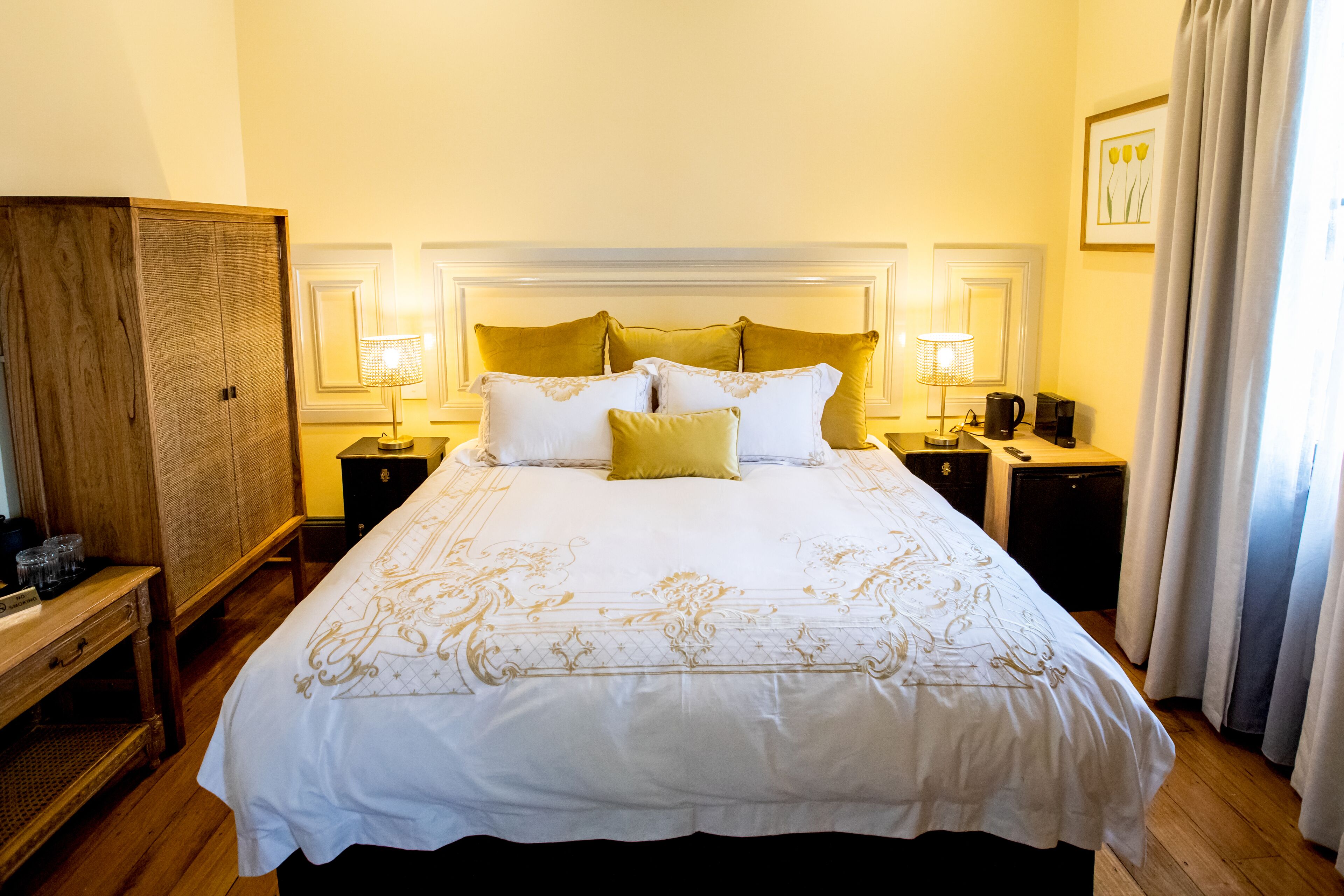 A neatly made bed with a white and gold bedspread, two bedside lamps, and a wooden wardrobe.