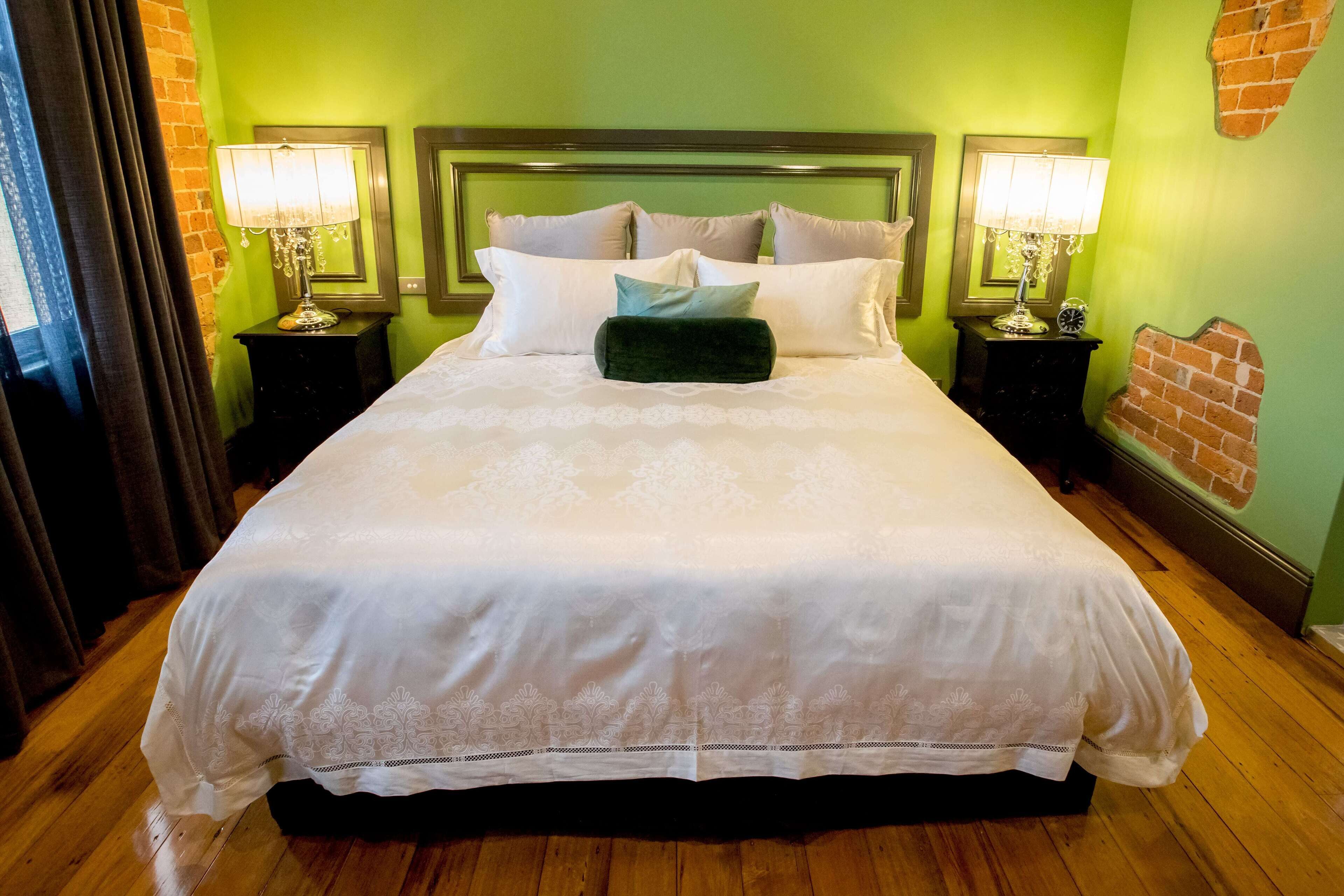 Un lit bien fait, avec du linge de lit blanc et un coussin vert, est encadré par deux tables de chevet sur lesquelles sont posées des lampes. La pièce est décorée de murs verts et le sol est recouvert de parquet.