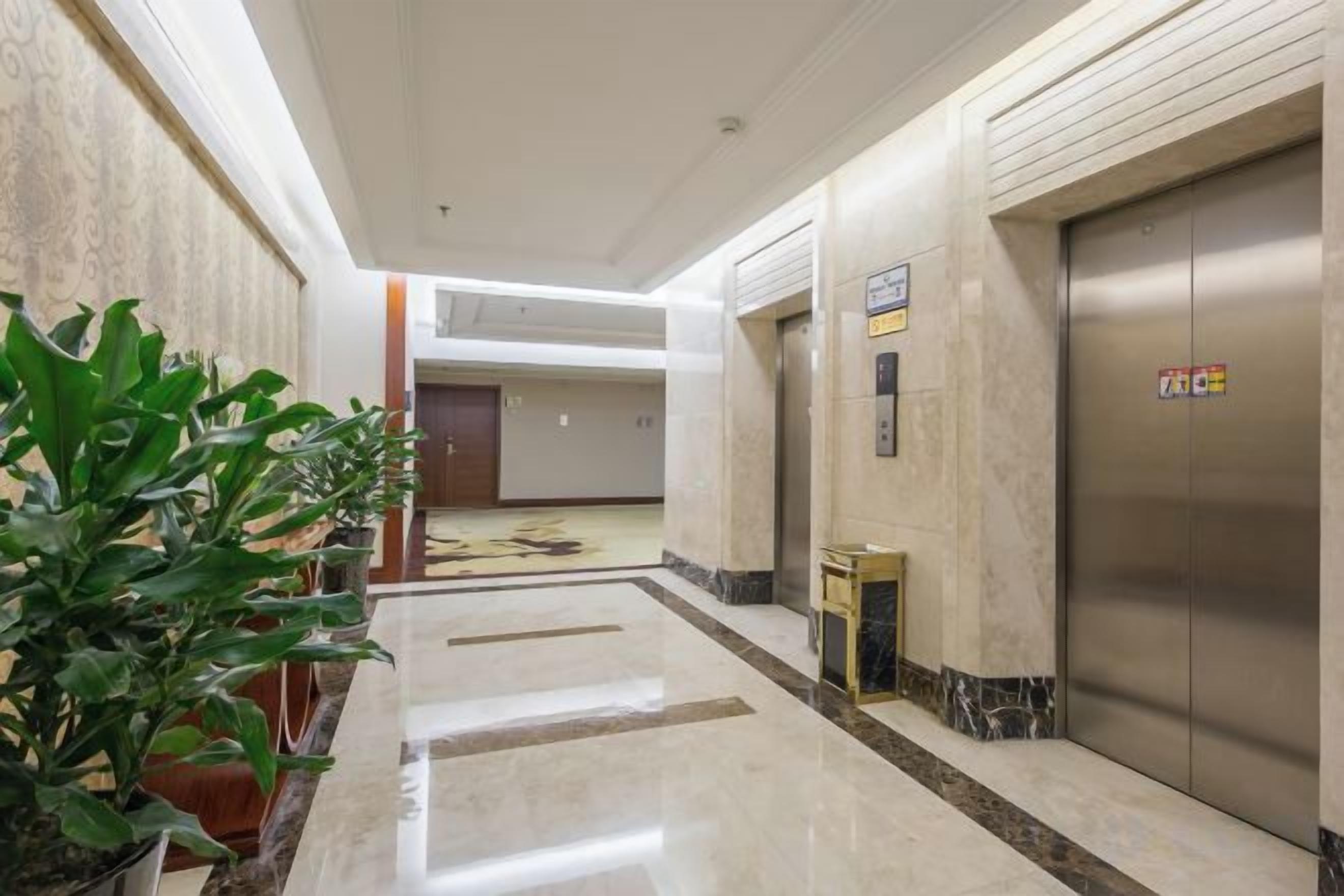 Un pasillo de hotel con piso de mármol, plantas en macetas, un mostrador de recepción y un ascensor.