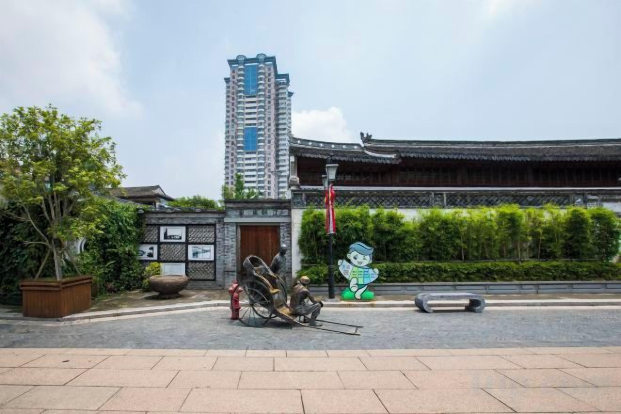 Un bâtiment traditionnel avec, en arrière-plan, un immeuble moderne de grande hauteur, une zone d’entrée pavée et une statue représentant un personnage.
