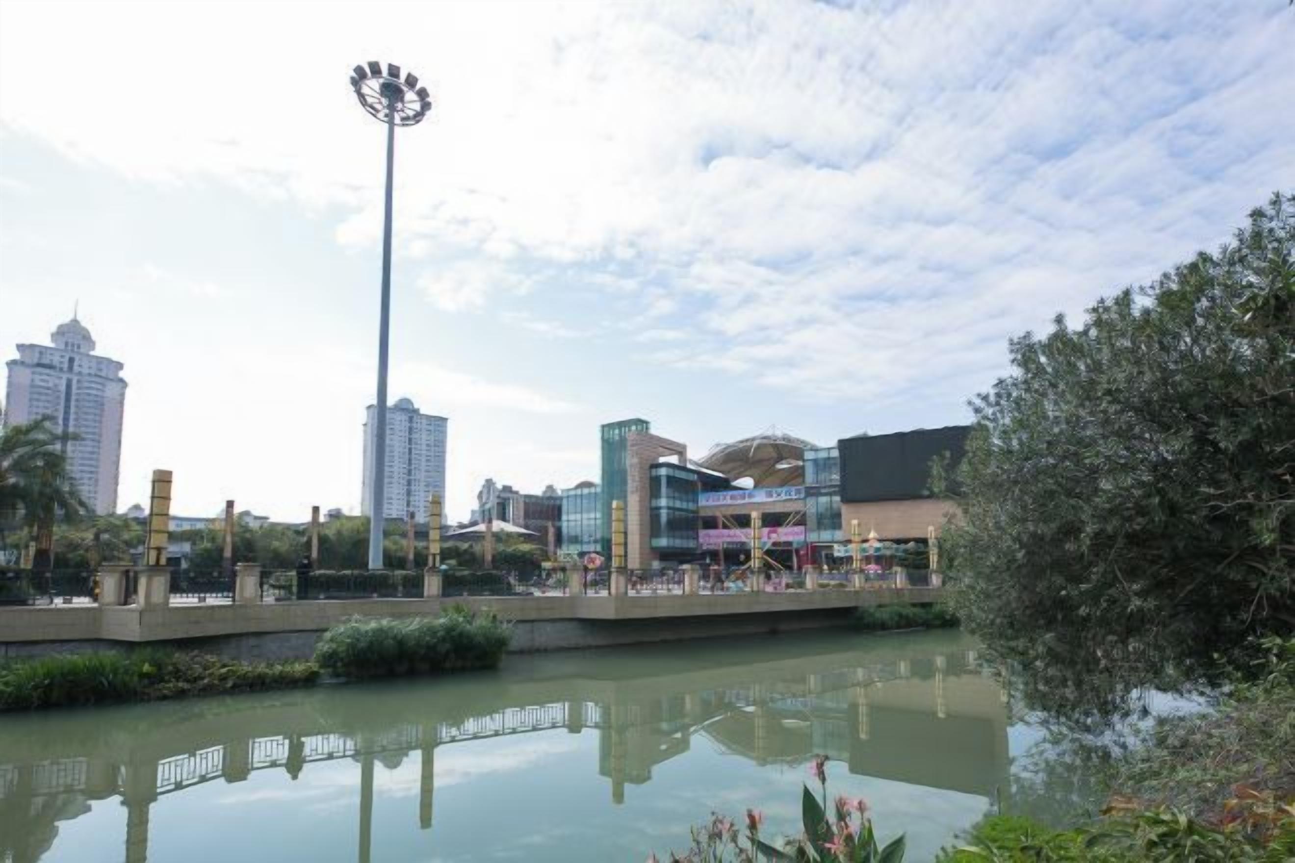 Un paisaje urbano con edificios modernos, una plaza central con una fuente y una masa de agua que refleja las estructuras.