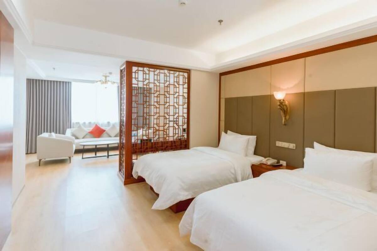 Hotellihuone, jossa on kaksi sänkyä, sohva, koristeellinen sermi ja seinään kiinnitetty valaisin.