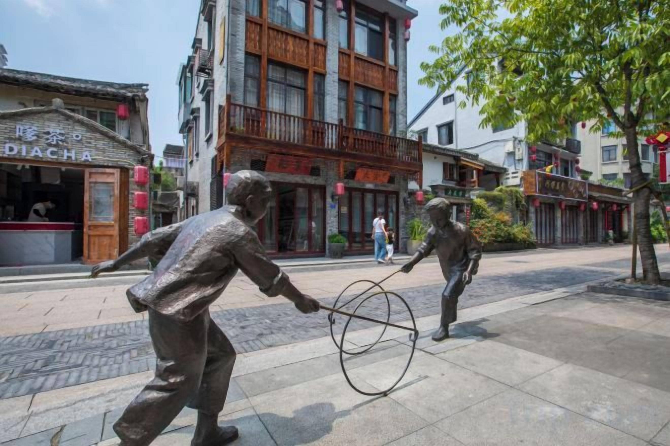 A street scene with a bronze statue of two figures playing a hoop, buildings with signage, and a pedestrian in the background.