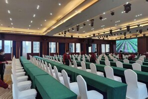 Un salón de conferencias con filas de mesas, sillas blancas y una pantalla grande al frente.