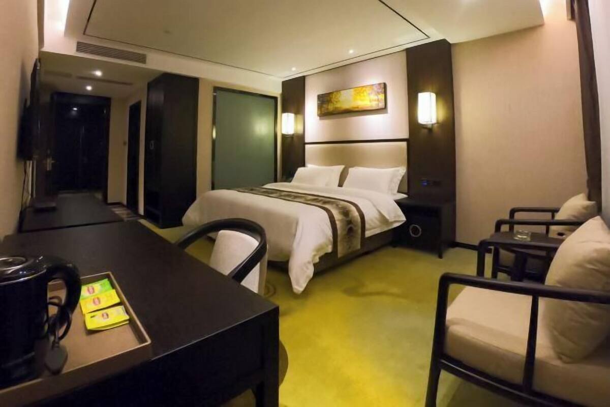 A hotel room with a bed, a desk with a kettle, a chair, a bedside table, and a painting on the wall.