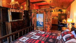A cozy cabin interior with a wooden interior, a bed with a quilt, a television, and a kitchen area visible through a door. - The Ole Pole Barn at Dry Creek Hideaways (Middle Valley)