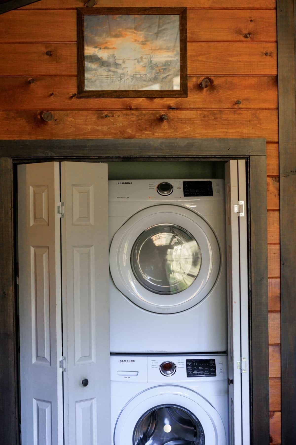 A front view of a laundry room with a Samsung washer and dryer, wooden walls, and a door with a painting above it.