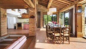 A spacious dining area with a brick fireplace, wooden beams, and a view of the outdoors. - Il Romitoio 12 (Castiglion Fiorentino)