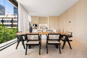 Een moderne eetruimte met een houten tafel en zwarte stoelen, een keuken op de achtergrond en uitzicht op de stad door grote ramen.
