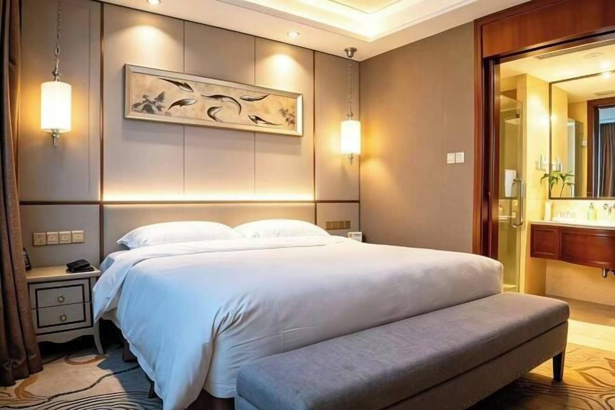 Ein Hotelzimmer mit einem großen Bett, Nachttischen, einer Bank, einem Wandbild und einem durch eine offene Tür einsehbaren Badezimmer.
