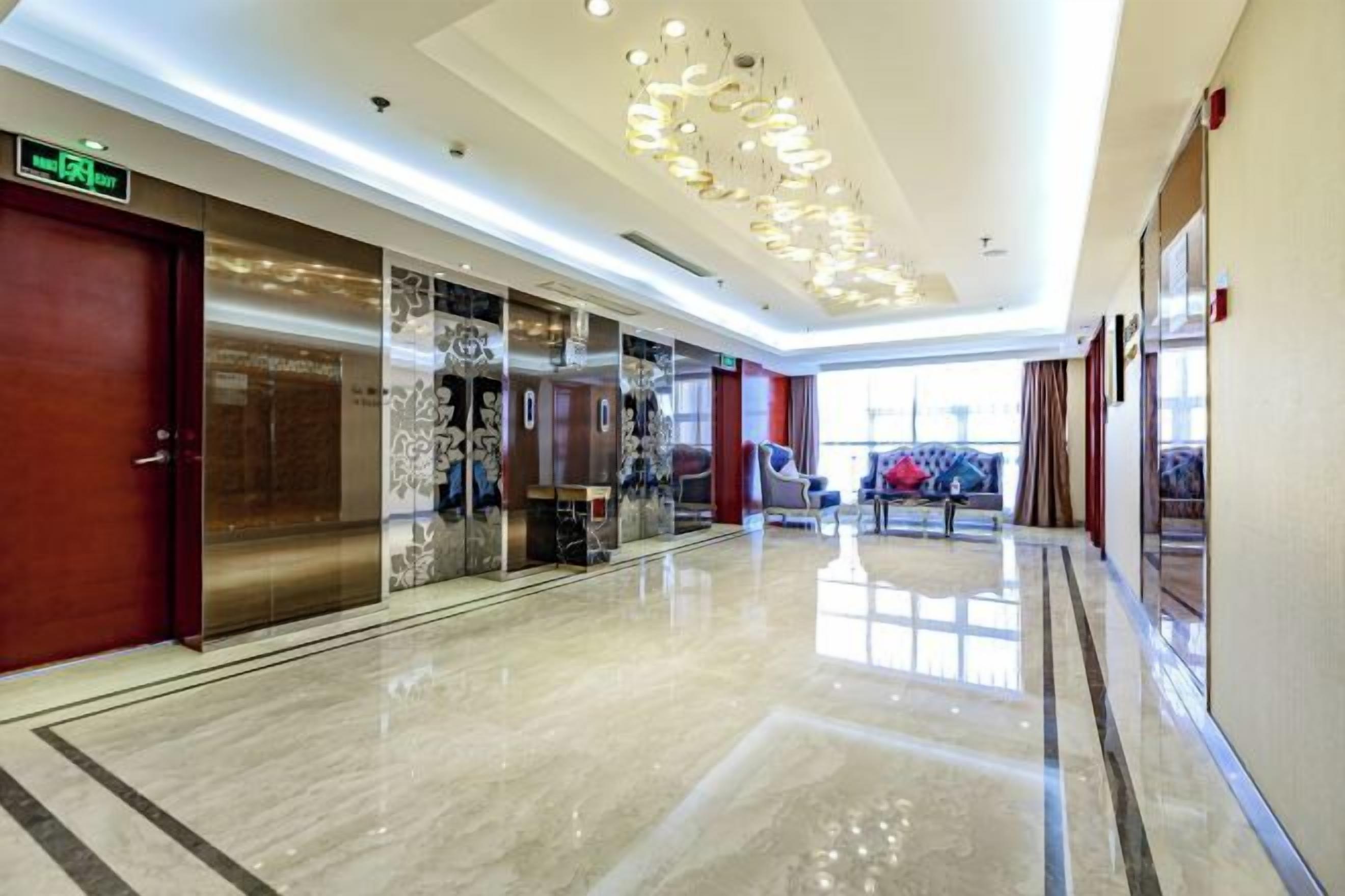 A hotel corridor with a polished floor, a modern chandelier, and a view of the outdoors through large windows.