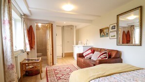 Un dormitorio con una cama, una silla de madera, una mesita, un sofá, un tapete rojo y beige, un armario de madera, un espejo y una ventana con cortinas.
