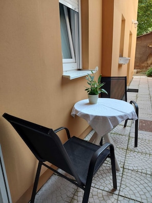 Ein kleiner Außenbereich mit Tisch und zwei Stühlen, einem Blumentopf und einem Fenster mit Jalousien.