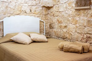 Una cama con estructura de metal blanco, ropa de cama color beige y un muro de piedra de fondo.