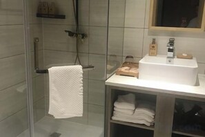 Une salle de bain avec une douche à parois de verre, un lavabo blanc avec un porte-savon et un porte-serviettes sur lequel est posée une serviette blanche.