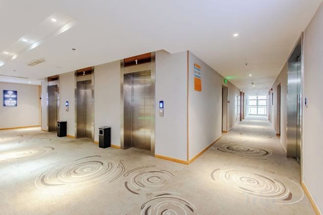 A hotel corridor with multiple elevators, a carpeted floor, and a long hallway.