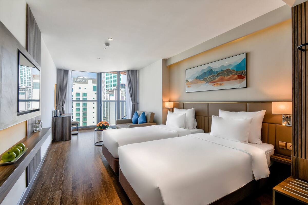 Una habitación de hotel moderna con dos camas, un ventanal con vistas a la ciudad, un cuadro en la pared y una mesita con un jarrón de flores.