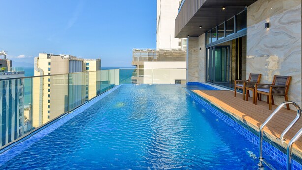Une piscine à débordement située sur le toit, avec une balustrade en verre, une terrasse en bois et un espace salon extérieur.