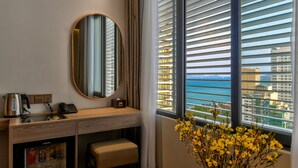 Une chambre d’hôtel avec un bureau en bois, un miroir rond, un téléphone, un petit réfrigérateur et une vue sur la mer grâce à de grandes fenêtres équipées de stores.