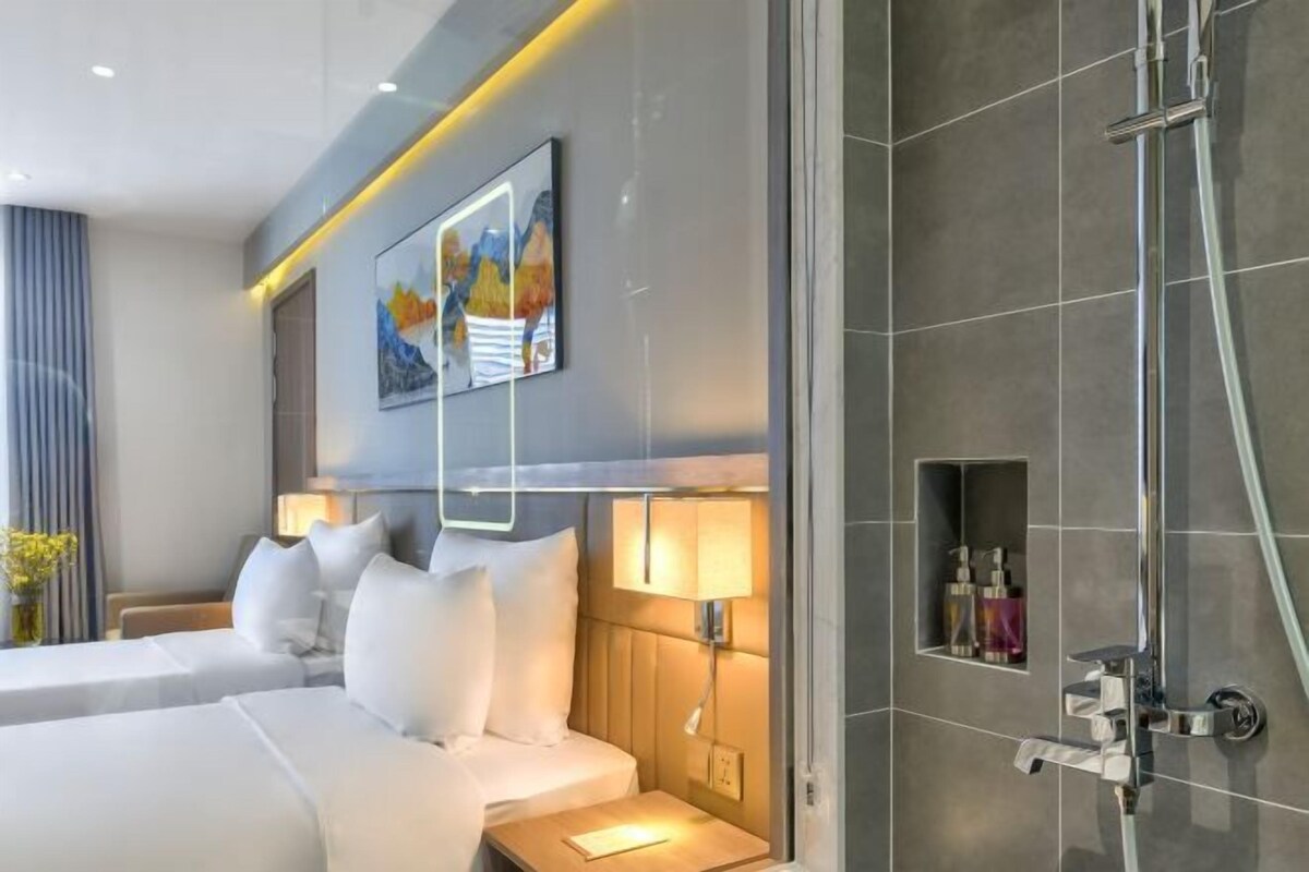A modern hotel room with two beds, a bedside table with a lamp, a shower area, and a framed artwork on the wall.