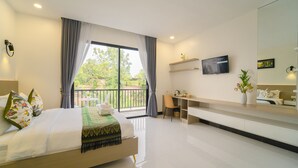 Habitación de hotel moderna con una cama grande, un escritorio, una silla, un televisor y un balcón con vistas a zonas verdes.