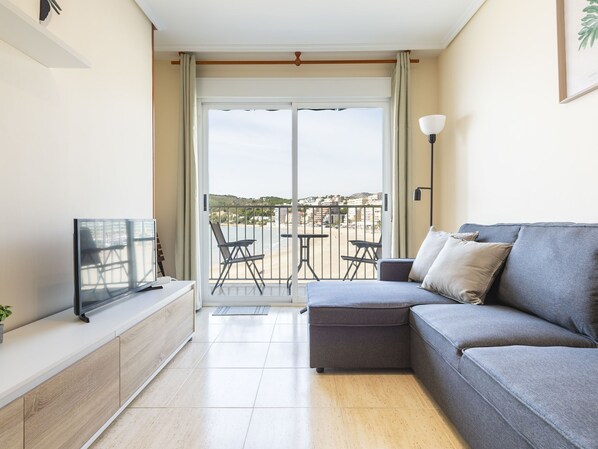 Una sala de estar moderna con un sofá gris, un televisor de pantalla plana y un balcón con vista a la playa.