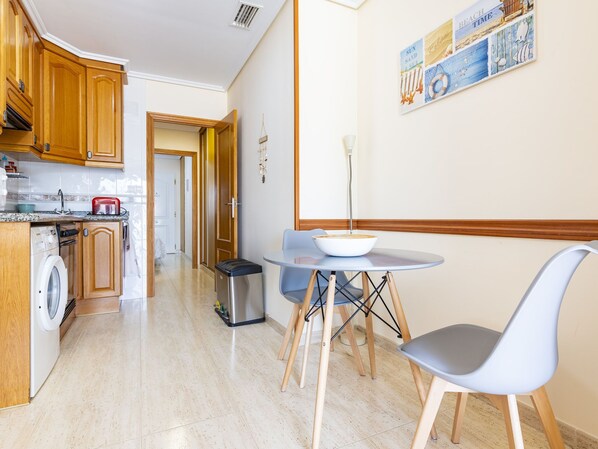 A modern kitchen with wooden cabinets, a washing machine, a trash bin, and a dining area with a table and chairs.
