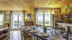 Zona pranzo con un tavolo lungo apparecchiato per sei persone, circondato da sedie di legno. Si vede l'esterno attraverso ampie finestre, ed è presente un quadro appeso al muro.