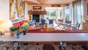 Un accogliente soggiorno con soffitto in legno, camino, un'ampia tela astratta e vista all'esterno tramite ampie finestre.