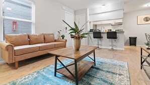 A modern living room with a leather sofa, a wooden coffee table, and a potted plant. A kitchen with a bar area is visible in the background. - Stylish 1BR Near UT Highland Evonify (Austin)