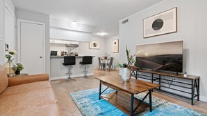 A modern living room with a sofa, a coffee table, and a TV. There is a kitchen in the background with a dining area. - Stylish 1BR Near UT Highland Evonify (Austin)