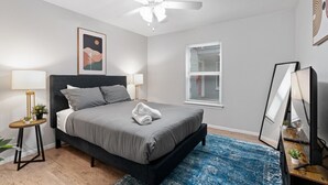 A bedroom with a bed, bedside lamps, a ceiling fan, a window with blinds, a framed picture on the wall, and a desk with a computer and plants.