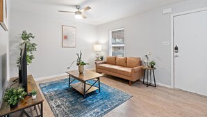 A living room with a brown sofa, a coffee table, a blue rug, a ceiling fan, a wall-mounted lamp, and a door with a keypad lock.