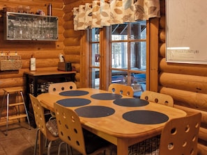 A wooden interior with a dining table, chairs, a bar area with glasses, and a window with a view.