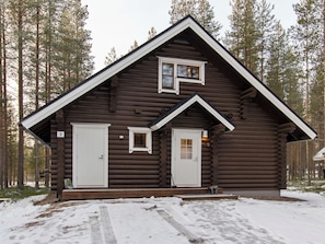 A log cabin with two doors and a small window, surrounded by a snowy landscape and tall trees.