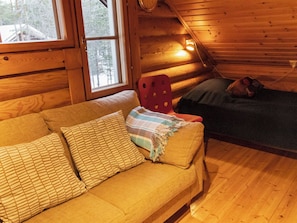 A cozy cabin interior with a wooden floor, a sofa with patterned cushions, a red chair, and a bed with a blanket.