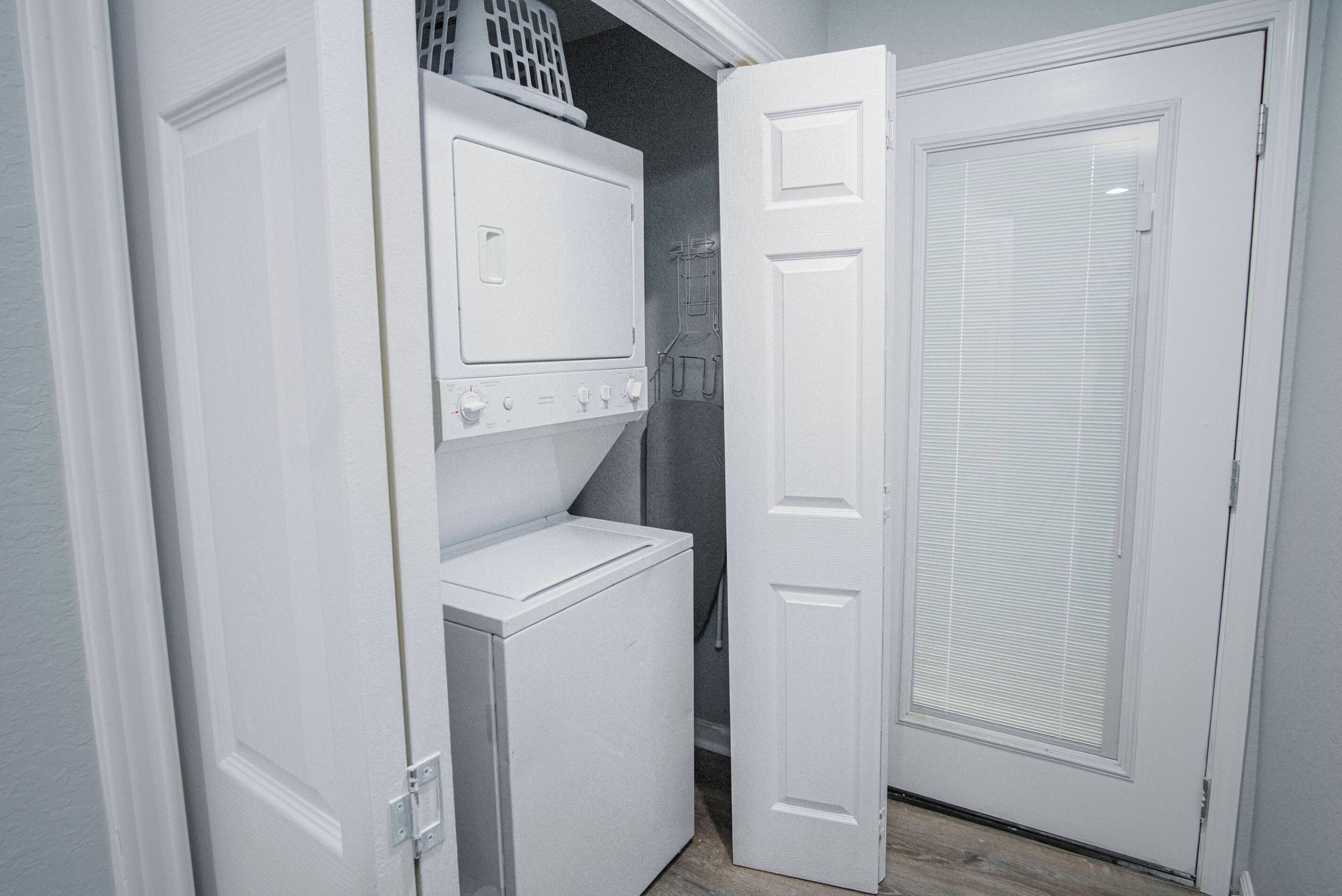Une buanderie équipée d’une machine à laver et d’un sèche-linge superposés, d’un panier à linge et d’une porte avec une fenêtre.