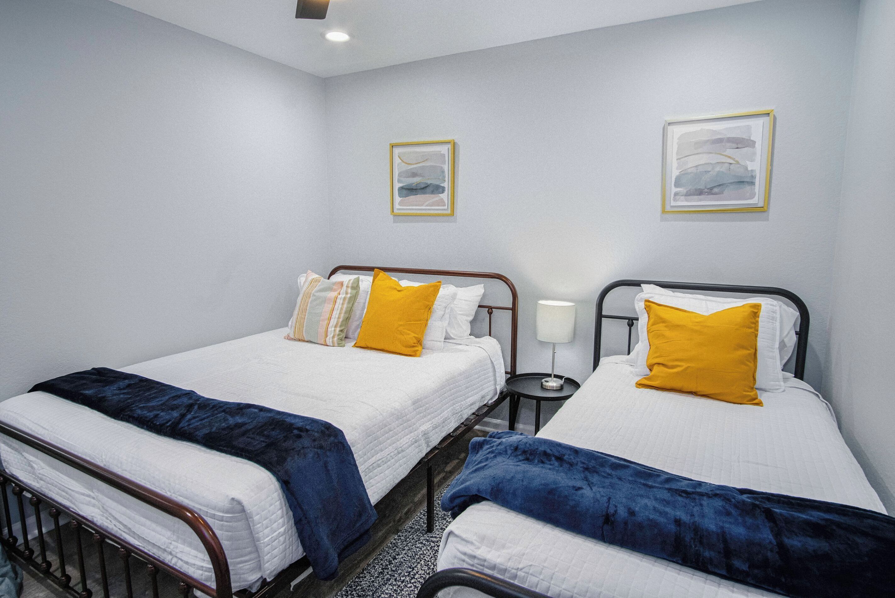 A hotel room with two beds, each with a white mattress and blue blankets. There are two bedside tables with lamps, and the walls are painted light grey.
