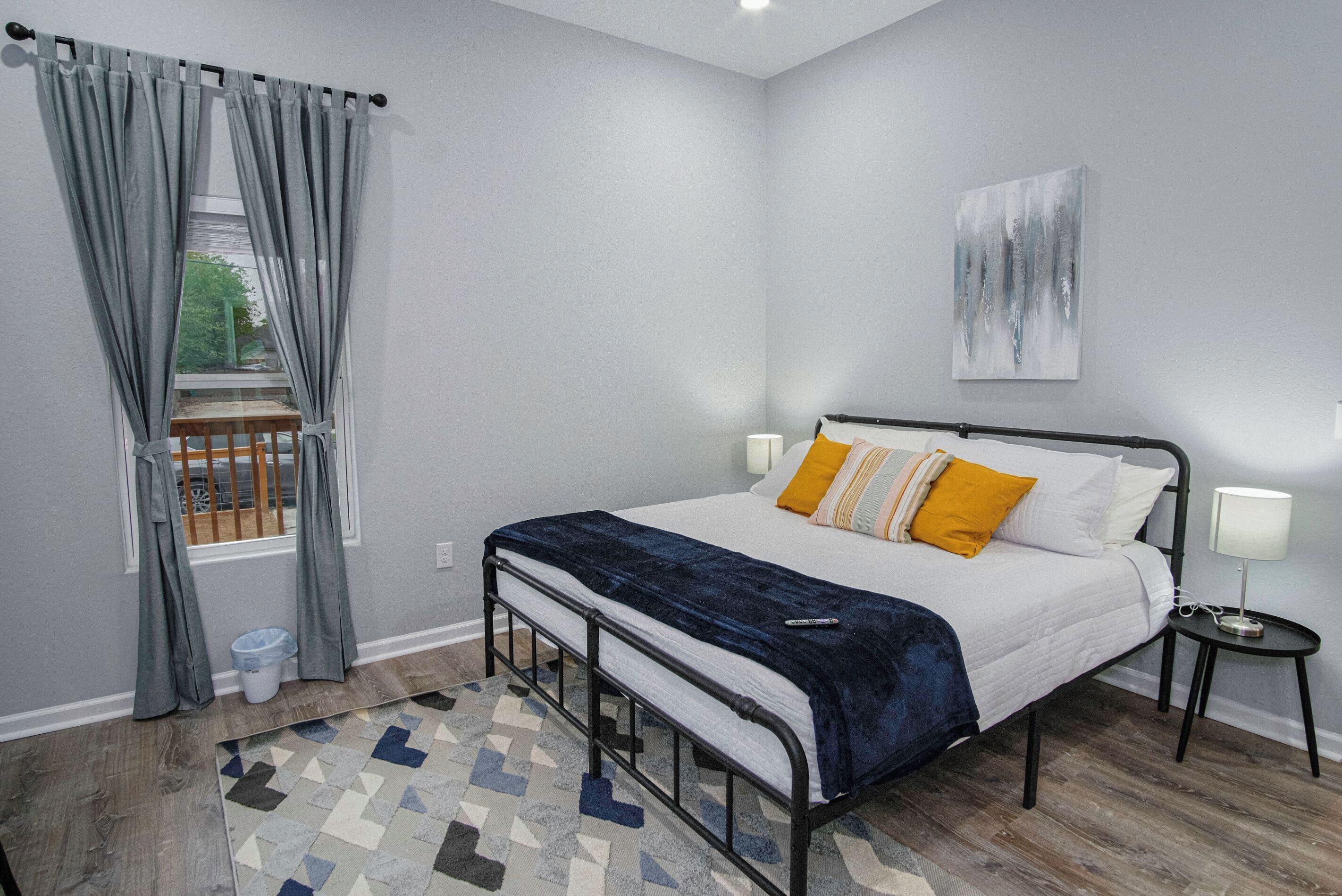 A bedroom with a bed, a window with curtains, a bedside table with a lamp, and a geometric-patterned rug.