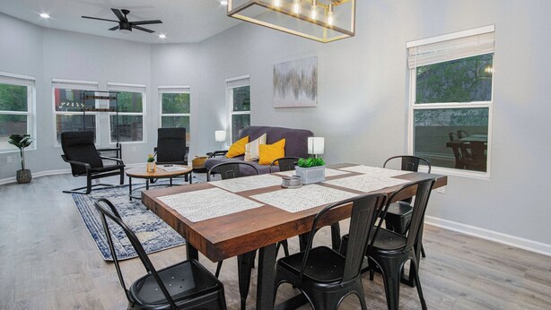 A spacious living room with a large wooden dining table, black chairs, a ceiling fan, and a modern chandelier.
