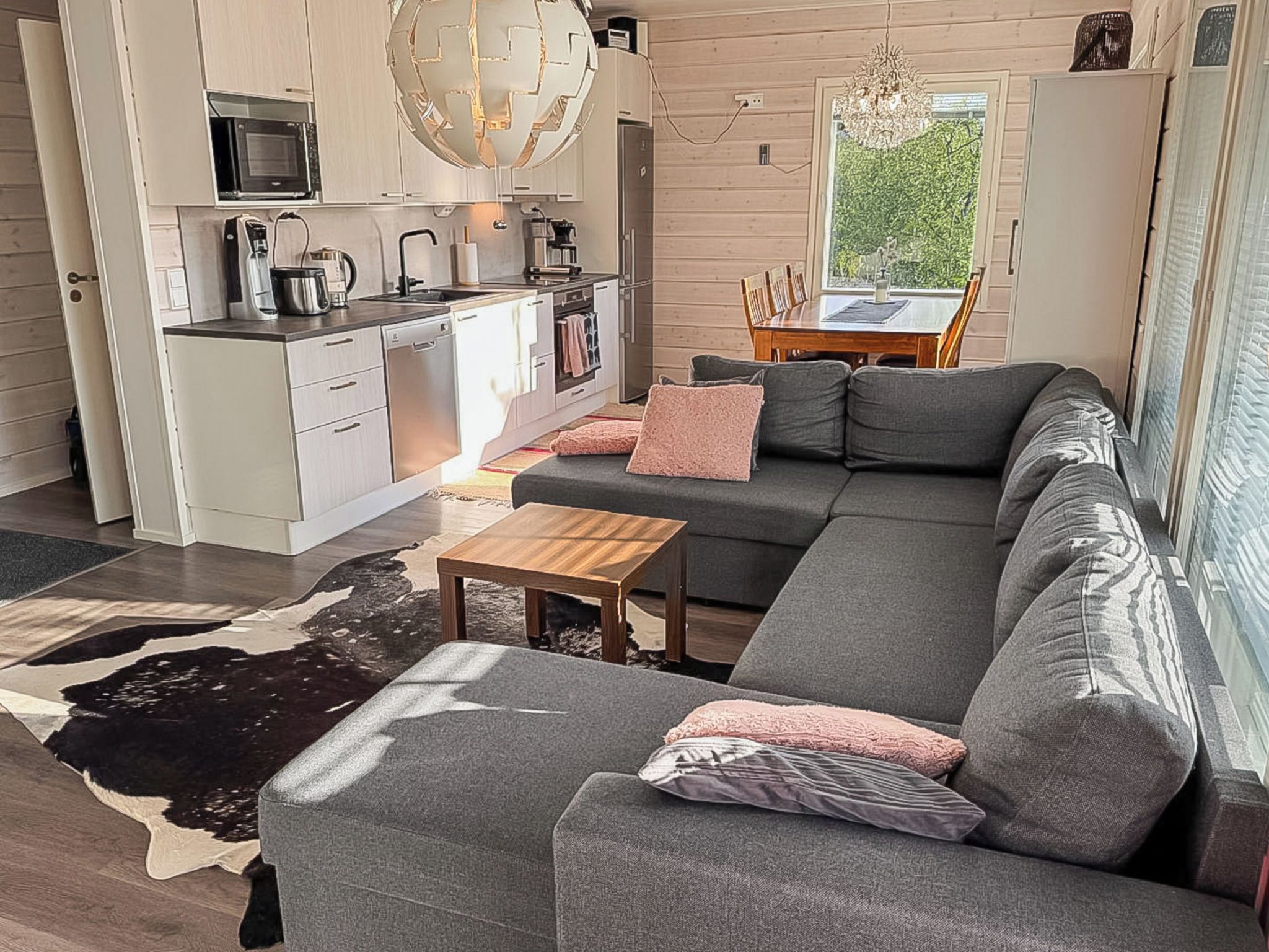 A modern living space with a grey sectional sofa, a wooden coffee table, a kitchen area with white cabinetry, and a dining table with chairs.