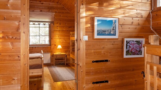 A wooden cabin interior with a bed, a table with a lamp, and two framed pictures on the wall.