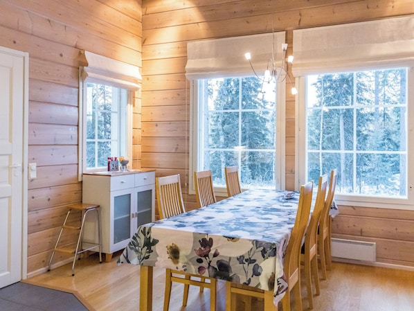 A wooden cabin interior with a dining table, chairs, a small kitchen area, and large windows with a snowy forest view.