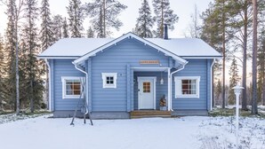 Een blauwe houten hut met een witte deur en ramen, met de tekst 'OUTA-KURSUUT', omringd door sneeuw en bomen.