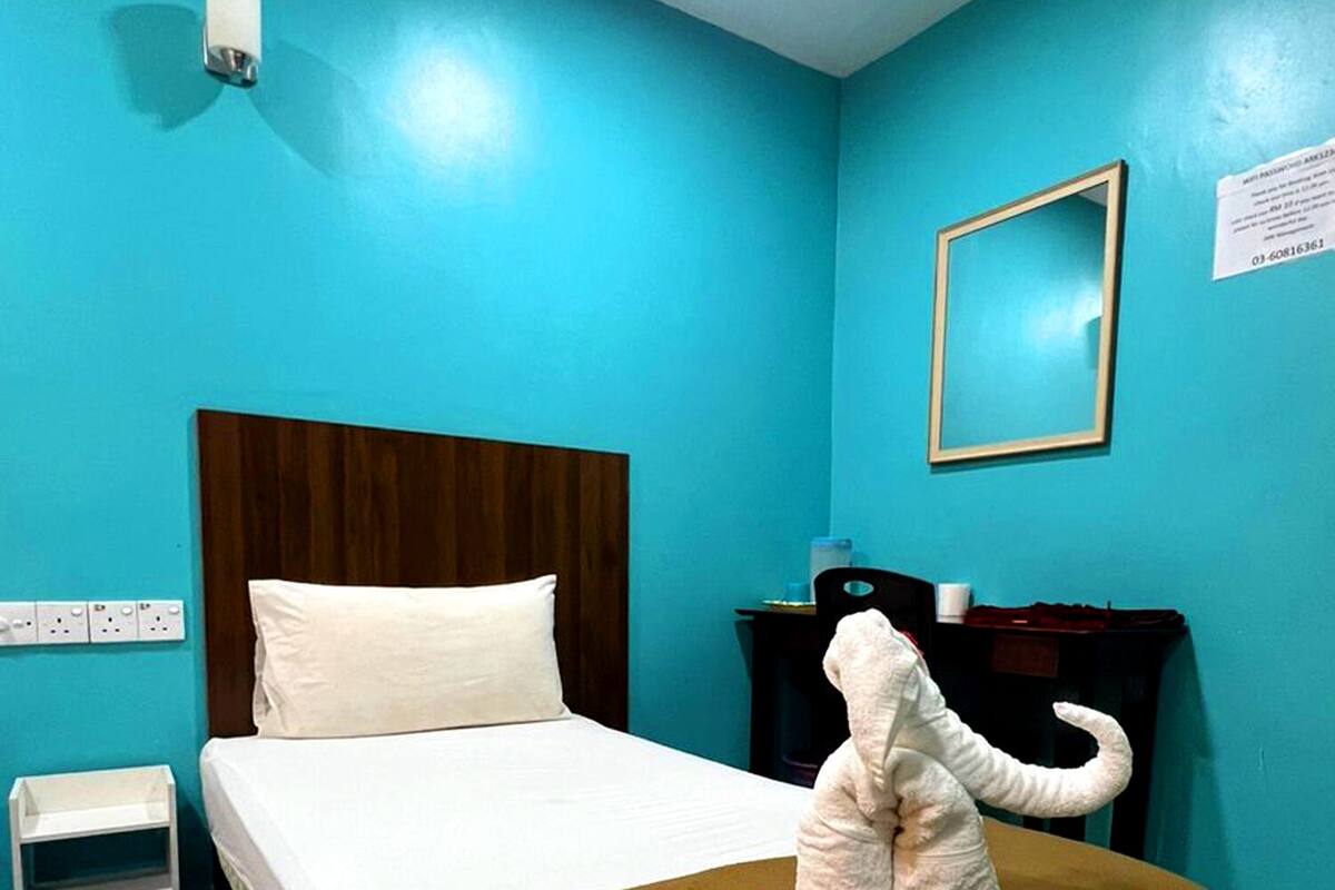 A hotel room with a bed, a nightstand, a wall-mounted light, a framed mirror, and a stuffed elephant toy on the bed.