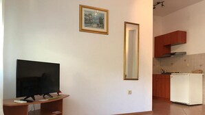 Uma sala de estar com televisão de tela plana sobre um suporte de madeira, um quadro na parede e um espaço de cozinha com armários e uma geladeira.