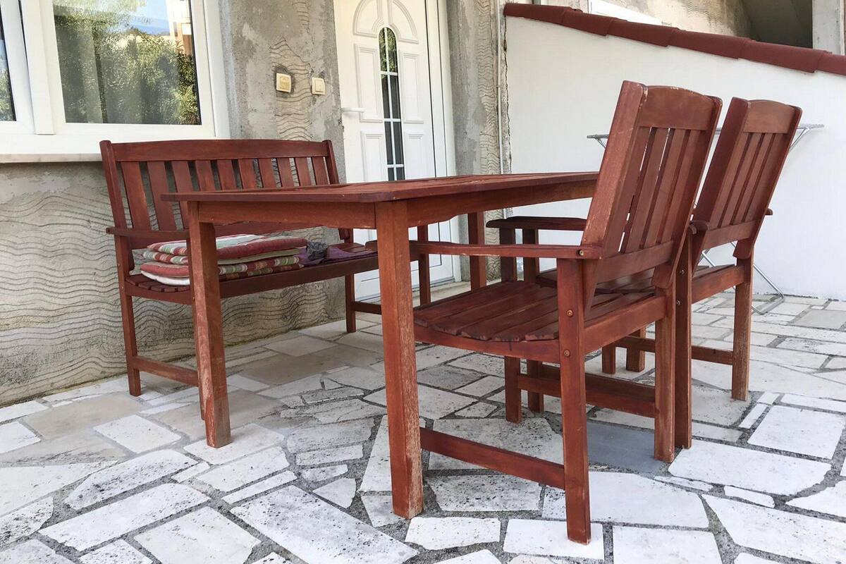 Une table et des chaises en bois sont disposées sur une terrasse carrelée en pierre, avec une porte et une fenêtre blanches en arrière-plan.
